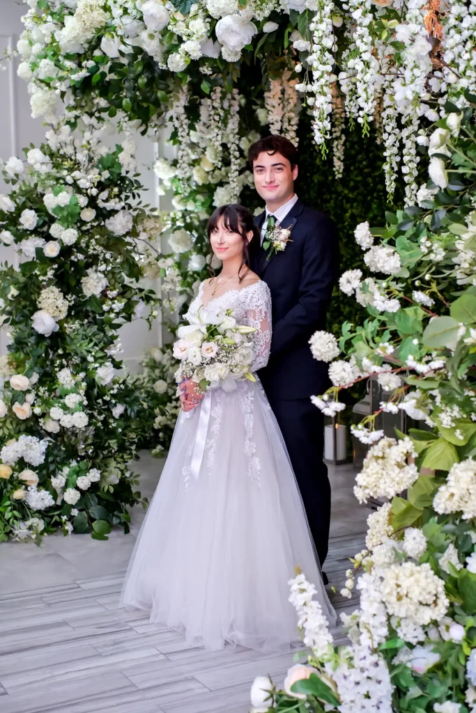 Romantic wedding portrait of a couple at Albertson Wedding Chapel — the bride in a sheer white gown holding a bouquet, standing before the chapel’s flower installation.